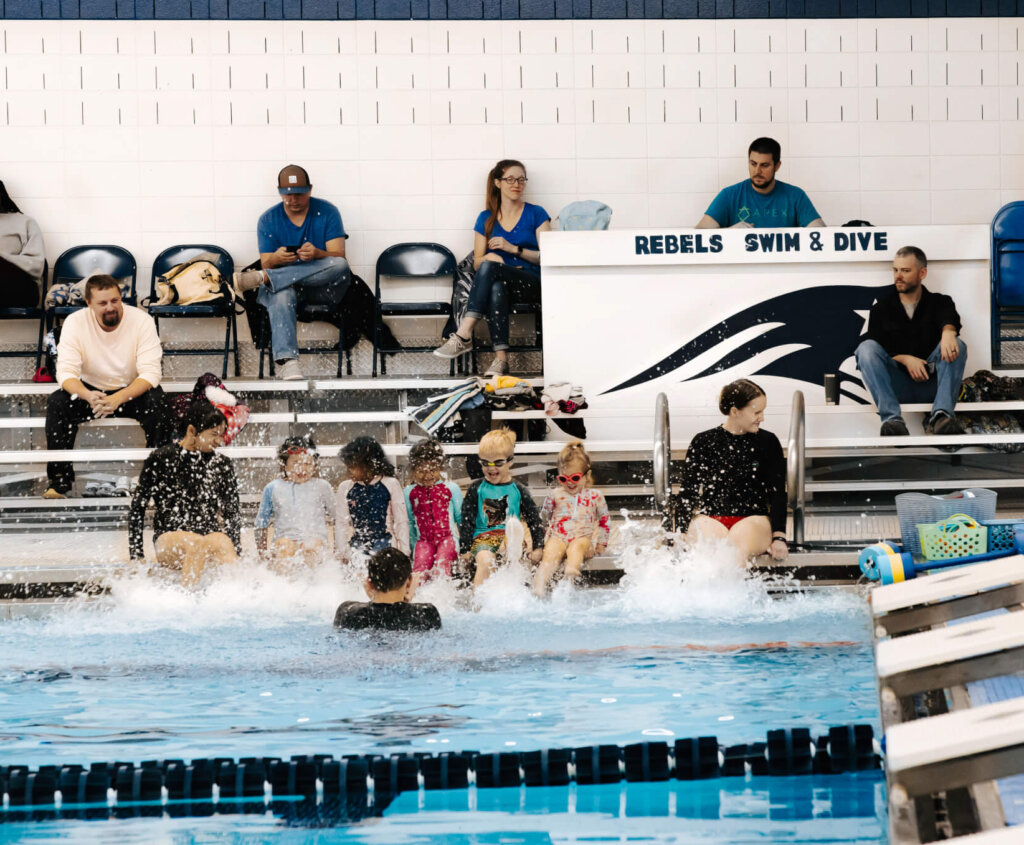Kids splashing in a pool at a swimming lesson. Adults in background