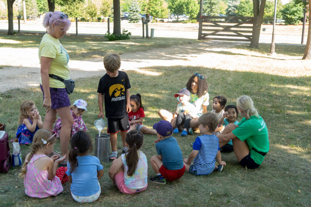 Kids gathered in a circle on a farm. One kid pouring milk to make ice cream. Adult watching on.